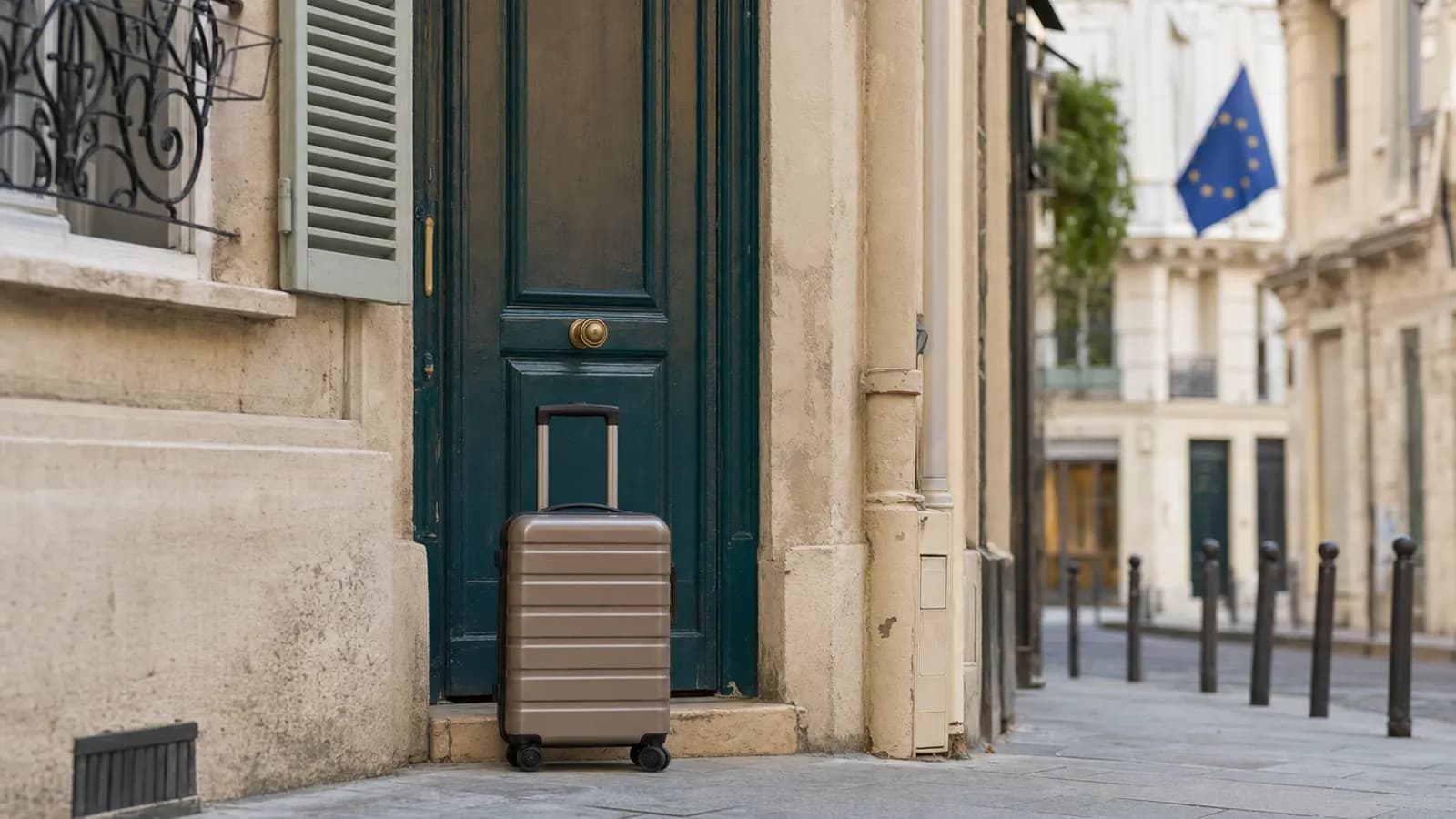 Valise à roulettes devant la porte d'un immeuble ancien, drapeau européen flottant au loin.