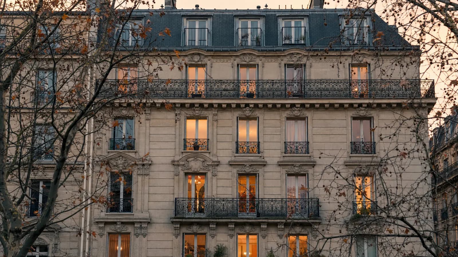 Façade d'immeuble haussmannien avec balcons en fer forgé, quelques fenêtres éclairées en fin de journée d'automne.