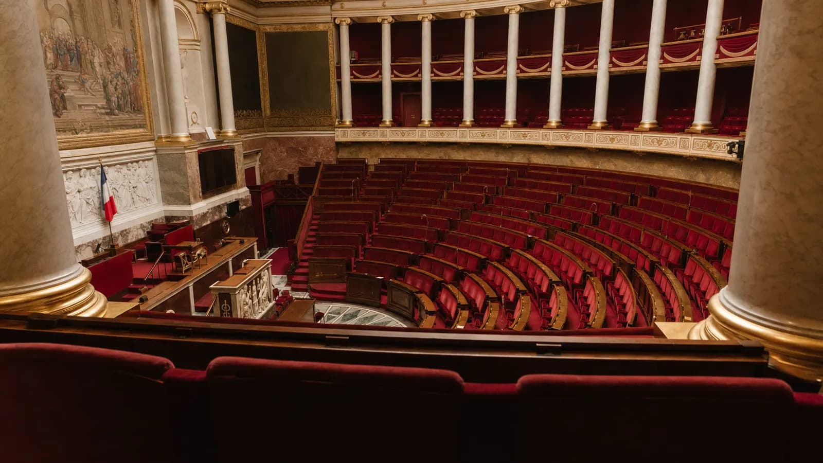 Hémicycle vide de l'Assemblée nationale vu des tribunes : sièges rouges en arc de cercle, colonnes de marbre, éclairage chaud.
